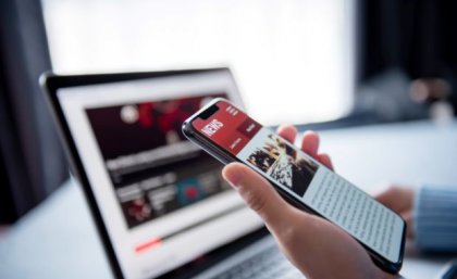 Image: Adobe hand holding a phone with screen on a news site in front of a laptop on a news web page